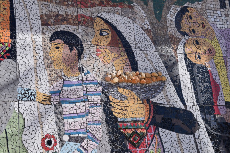 A close up of the memorial mosaic, focused on a woman wearing traditional Palestinian dress and white headscarf and cradling a basket of rocks, with a young boy pictured beside her.